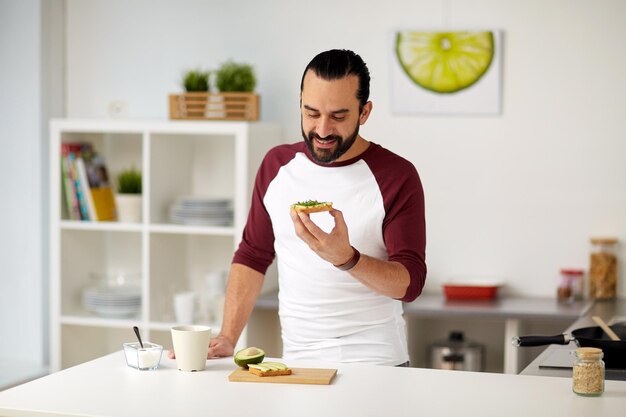 ベジタリアン料理、健康的な食事、人々、ダイエットのコンセプト – 自宅のキッチンで朝食にアボカドサンドイッチを持つ男性