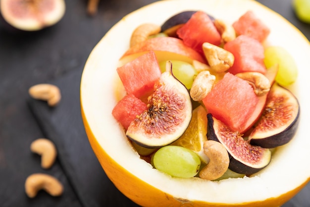 Vegetarian fruit salad of watermelon, grapes, figs, pear, orange, cashew  on slate board on a black concrete background. Side view, close up, selective focus.