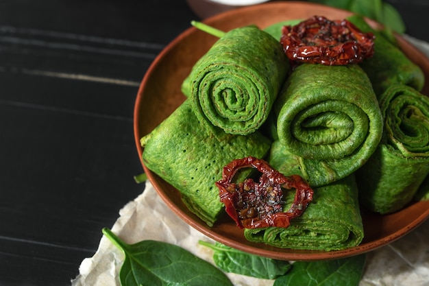 Vegetarian green pancakes decorated with dried tomatoes. Spinach pancakes rolls on a brown plate on dark wooden desk.