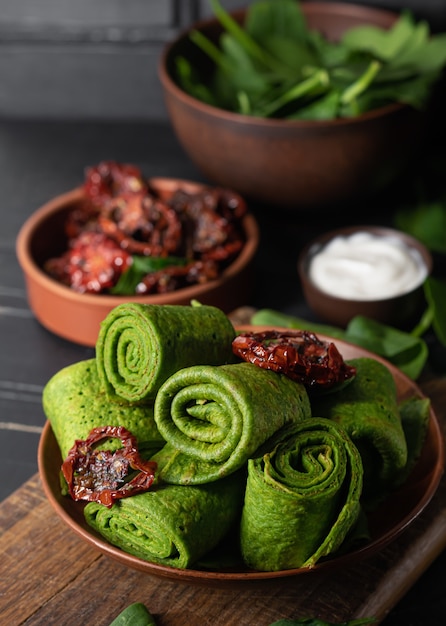 Vegetarian green pancakes decorated with dried tomatoes. Spinach pancakes rolls in a brown plate on dark wooden desk.