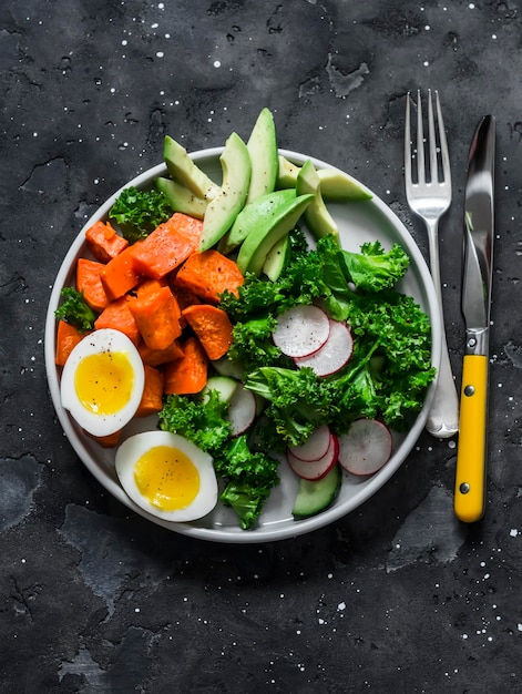 Vegetarian lunch boiled sweet potatoes egg kale avocado radish cucumber salad on a dark background top view