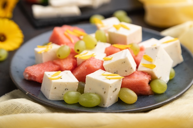 Vegetarian salad with watermelon, feta cheese, and grapes on blue ceramic plate on black concrete background and yellow linen textile. Side view, close up, selective focus.