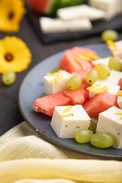 Vegetarian salad with watermelon, feta cheese, and grapes on blue ceramic plate on black concrete background and yellow linen textile. Side view, close up, selective focus.