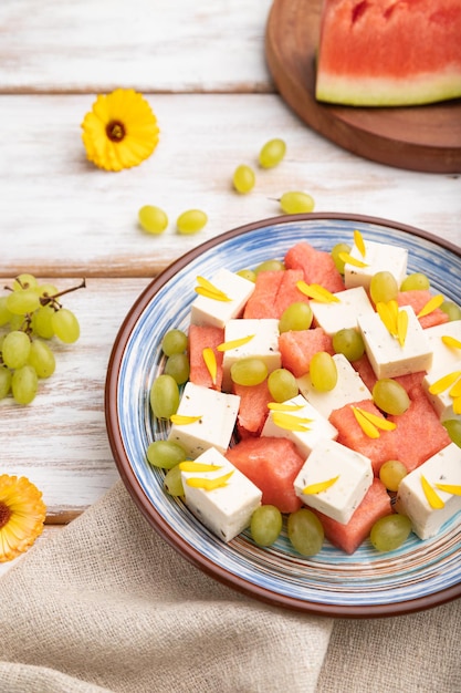 Insalata vegetariana con anguria, feta e uva su piatto di ceramica blu su fondo di legno bianco e tessuto di lino. vista laterale, primo piano, messa a fuoco selettiva.