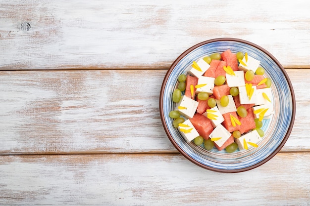 Vegetarian salad with watermelon feta cheese and grapes on blue ceramic plate on white wooden background top view copy space