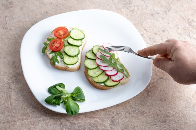 Vegetarian sandwiches with cottage cheese, cucumbers, tomatoes and radishes on a white plate. A hand holds a fork over a white plate with sandwiches.