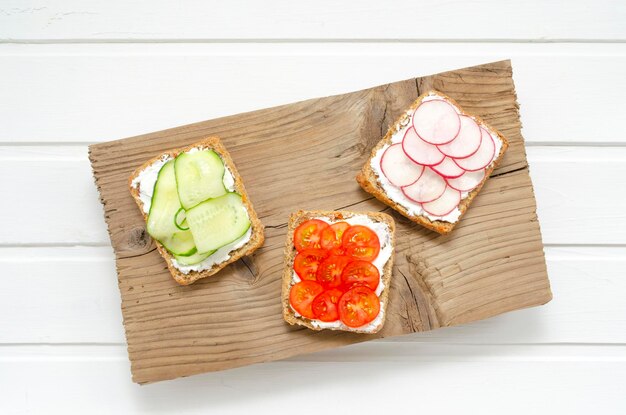 Vegetarian sandwiches with cream cheese fresh cucumber radish tomatoes on a board on a white wooden