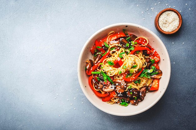 Vegetarian stir fry noodles with vegetables paprika mushrooms chives and sesame seeds in bowl Gray table background top view copy space