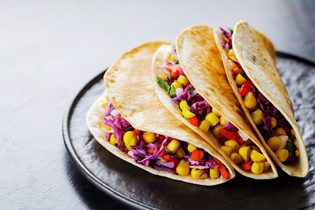 Vegetarian tacos with various vegetables, guacamole and lime on dark background. Tacos with sweet corn, purple cabbage and tomatoes on a black plate. Close-up