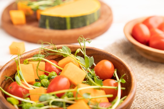 Vegetarian vegetable salad of tomatoes, pumpkin, microgreen pea sprouts on white wooden background and linen textile. Side view, close up, selective focus.