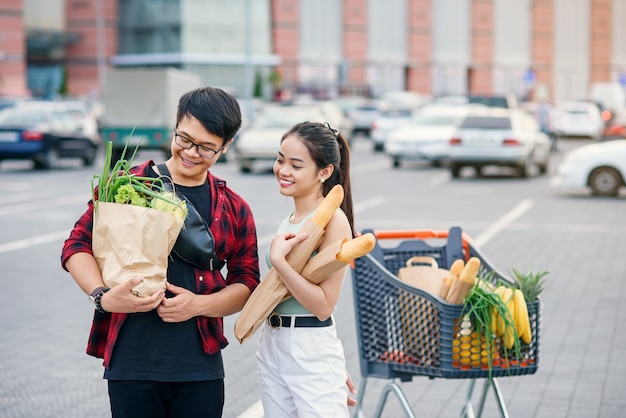 Vegetarian vietnamese couple hold paper eco bags with organic healthy food in hands while standing near store mall. Happy family shopping on weekend.