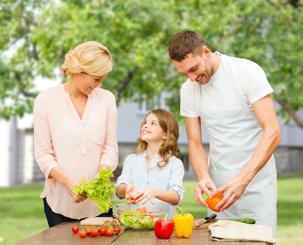 vegetarisch eten, culinair, geluk en mensenconcept - gelukkige familie die groentesalade kookt voor het diner over de achtergrond van het zomerhuis