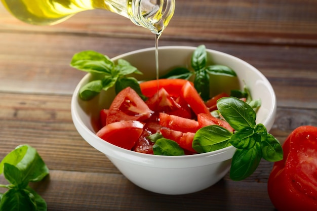Vegetarisch eten. Verse rode tomaten worden gesneden in een witte keramische plaat met basilicumkruid en olijfolie op een houten tafel.