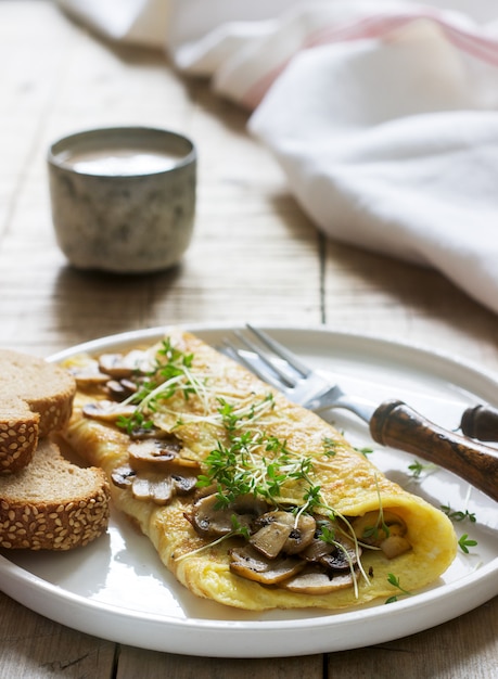 Vegetarisch ontbijt, omelet met champignons en tuinkers, geserveerd met roggebrood en koffie met melk. Rustieke stijl.