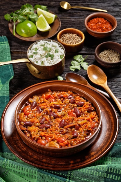 Vegetarische chili met bruine bonen en linzen in aardewerken kom op een donkere houten tafel met rijst en kruiden, verticale weergave van bovenaf