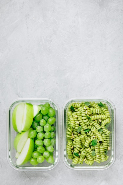 Vegetarische maaltijdbereidingscontainers groene pesto pasta met basilicum en pijnboompitten appel en druif als dessert bovenaanzicht kopieerruimte