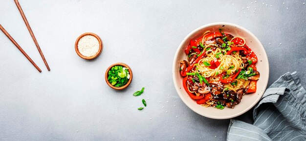 Vegetarische roerbaknoedels met groenten paprika champignons bieslook en sesamzaadjes in kom Grijze tafel achtergrond bovenaanzicht kopie ruimte