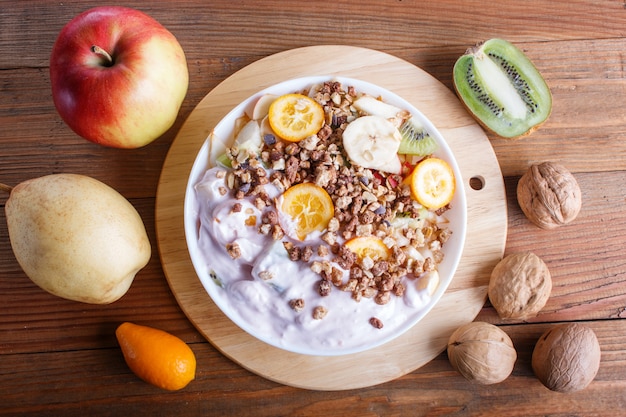 Vegetarische salade van bananen, appels, peren, kumquats, kiwi met muesli en yoghurt op bruin houten tafel.