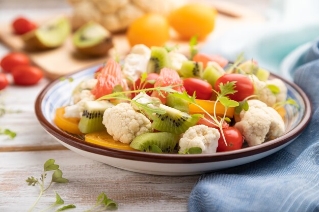 Vegetarische salade van bloemkoolkool, kiwi, tomaten, microgreenspruiten op witte houten achtergrond en blauw linnentextiel. zijaanzicht