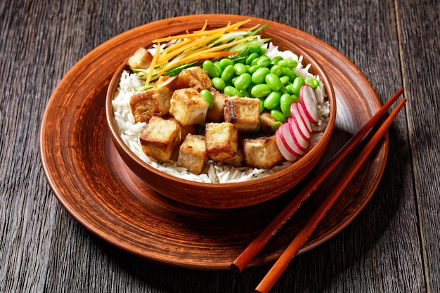 Vegetarische tofu poke bowl met basmati rijst edamame bonen radijs dun gesneden komkommer en wortelen salade in een klei kom met stokjes horizontale weergave van bovenaf