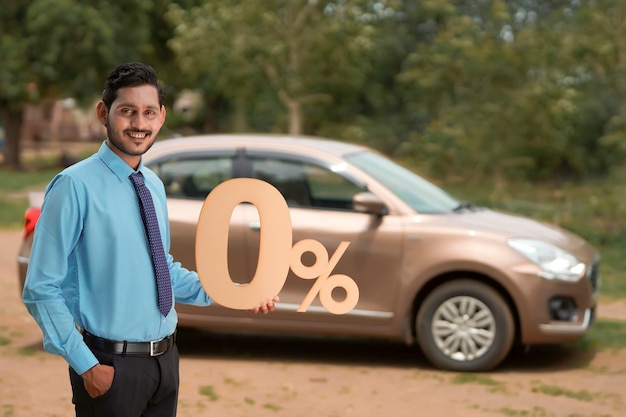 Vehicle loan concept : Young indian banker or financier showing zero percent sign symbol on new vehicle