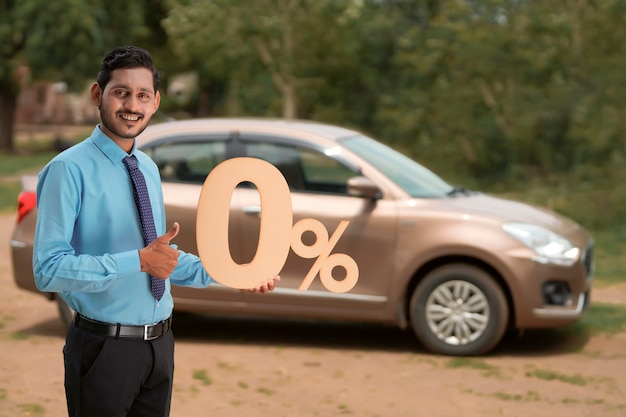 Vehicle loan concept : Young indian banker or financier showing zero percent sign symbol on new vehicle