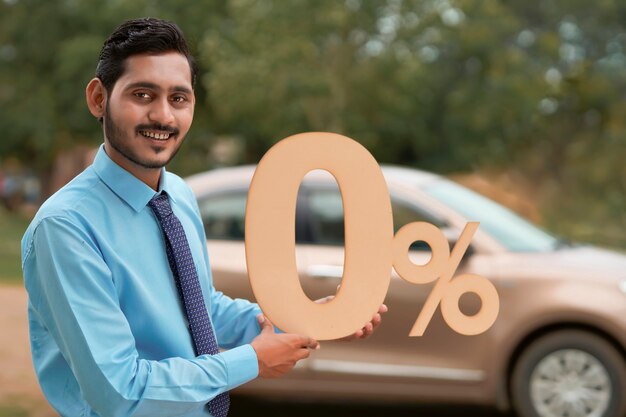 Vehicle loan concept : Young indian banker or financier showing zero percent sign symbol on new vehicle