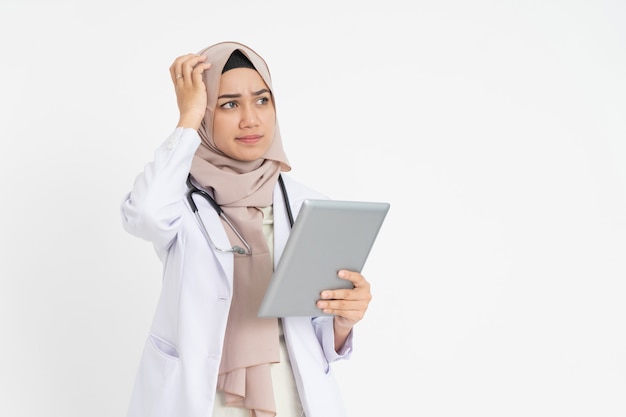 Veiled female doctor wearing white suit uniform confused while looking at pad screen with hand scrat...