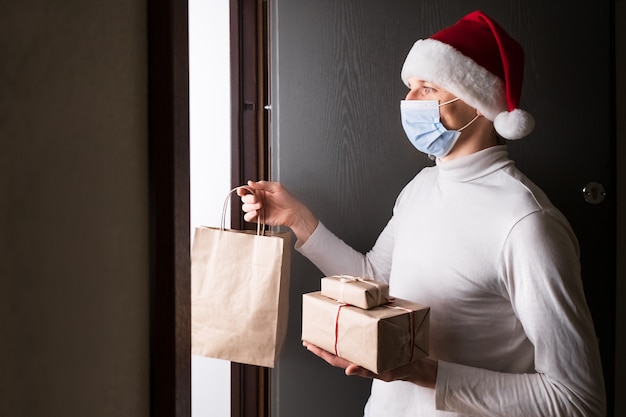 Veilige kerst- en nieuwjaarslevering, man-koerier in rode kerstmuts en beschermend masker tijdens coronavirus-quarantaine. Cadeautjes en thuis