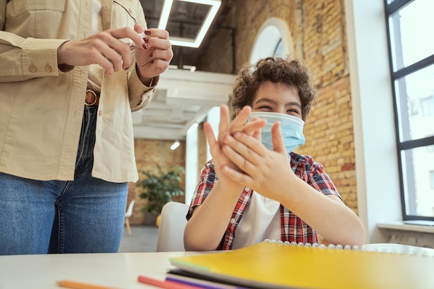 Veiligheid is belangrijk speelse kleine schooljongen die een beschermend masker draagt dat zijn handen schoonmaakt vrouw