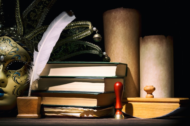 Venetian mask with old inkwell, feather, quill, scrolls, books and seal on wooden table. Vintage still life.