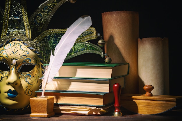 Venetian mask with old inkwell, feather, quill, scrolls, books and seal on wooden table.