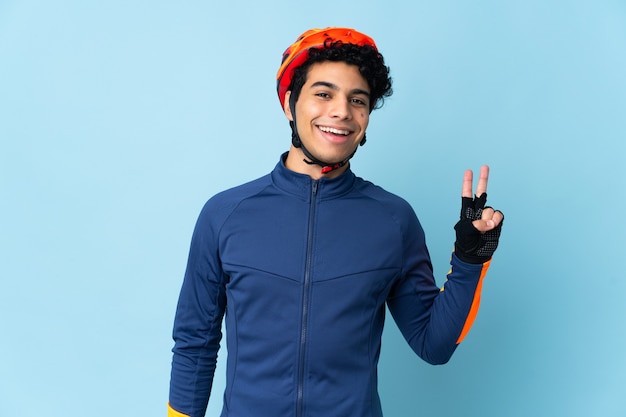 Venezuelan cyclist man isolated on blue wall smiling and showing victory sign