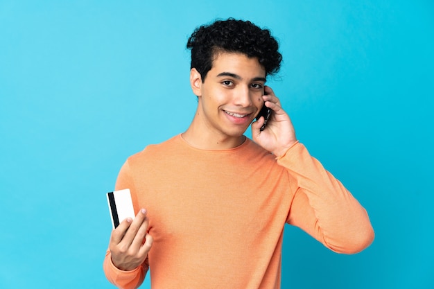 Venezuelan man isolated on blue keeping a conversation with the mobile phone and holding a credit card