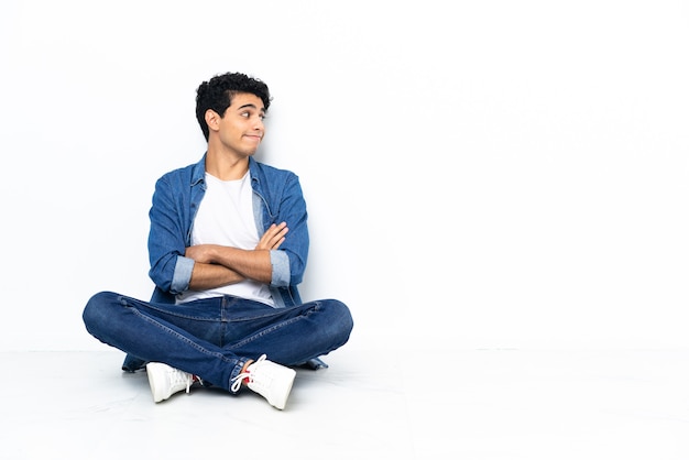 Venezuelan man sitting on the floor making doubts gesture while lifting the shoulders