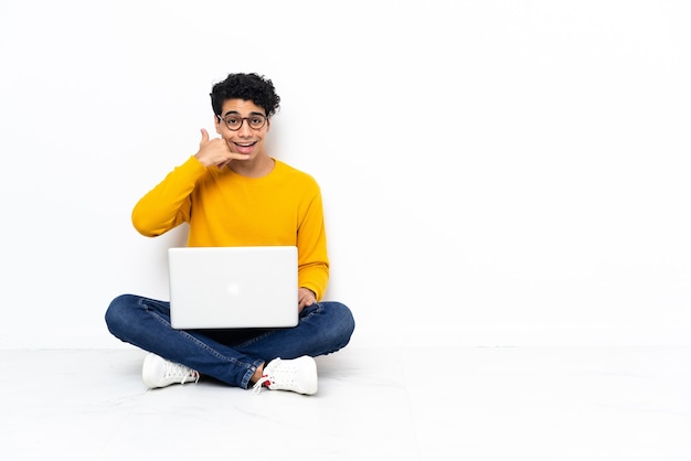 Venezuelan man sitting on the floor with laptop making phone gesture. Call me back sign