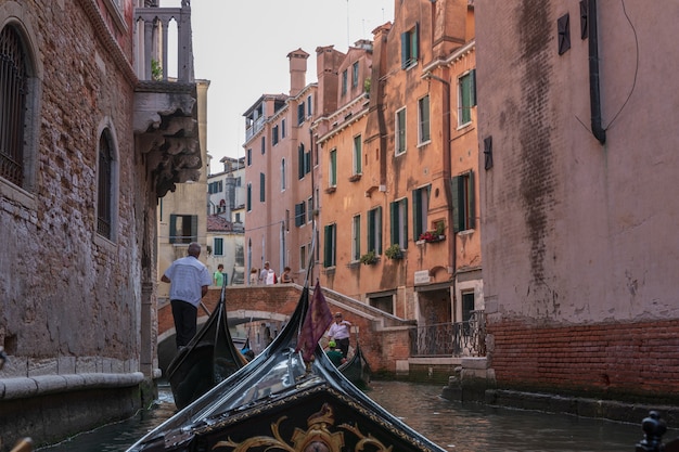 Venice, Italy - July 2, 2018: Panoramic view of Venice narrow canal with historical buildings and gondolas from other gondola. People relax in gondola. Summer sunny day and sunset sky
