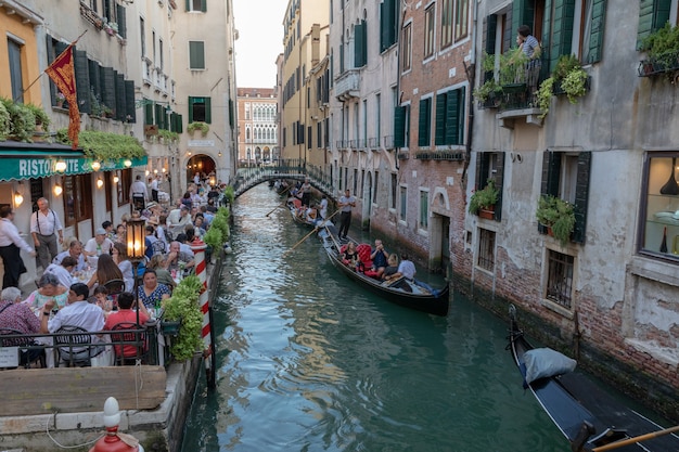 Venice, Italy - June 30, 2018: Panoramic view of Venice canal with historical buildings and gondolas from bridge. People relax and eating in restaurant. Landscape of summer evening day
