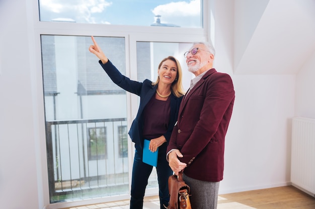 Venster tonen. Blondharige aantrekkelijke makelaar draagt stijlvolle elegante blouse met mooi groot raam in modern huis