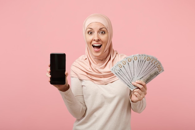 Verbaasd Arabisch moslimmeisje in hijab lichte kleding poseren geïsoleerd op roze achtergrond. Mensen religieuze levensstijl concept. Bespotten kopie ruimte. Houd mobiele telefoon vast met leeg leeg scherm, fan van contant geld.