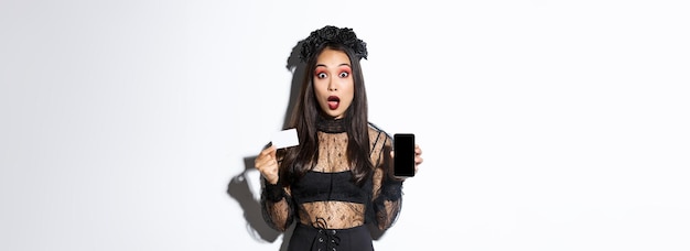 Verbaasd Aziatisch meisje in heksenkostuum dat creditcard met open mond van het mobiele telefoonscherm toont