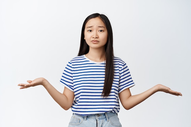 Verbaasd Aziatisch meisje weet niet wat ze moet doen, spreidt handen zijwaarts en doet verward schouderophalend gebaar, zonder idee op wit