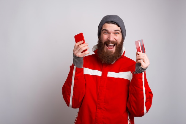 Verbaasd bebaarde hipster man met winterkleren en met smartphone en creditcard