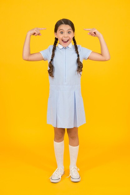 Verbaasd en blij. kind in vintage stijl wijzend op haar. ouderwets. kindermode. gelukkig kind bij gele muur. mode en schoonheid. jeugd geluk. klein meisje in schooluniform. retro-look.