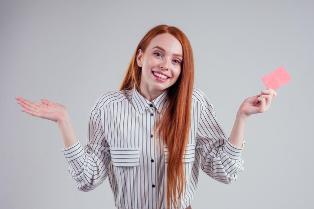 Verbaasd en verbaasd aantrekkelijke roodharige kaukasische zakenvrouw dragen gestreept shirt met creditcard studio witte achtergrond zwarte vrijdag saleguilty blik ik weet het niet