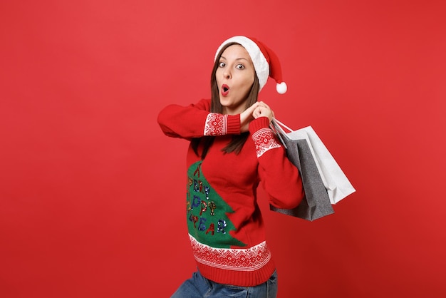 Verbaasd jong Santa-meisje dat de mond wijd open houdt en er verrast uitziet met pakketten met aankopen na het winkelen geïsoleerd op rode achtergrond. Gelukkig Nieuwjaar 2019 viering vakantie partij concept.