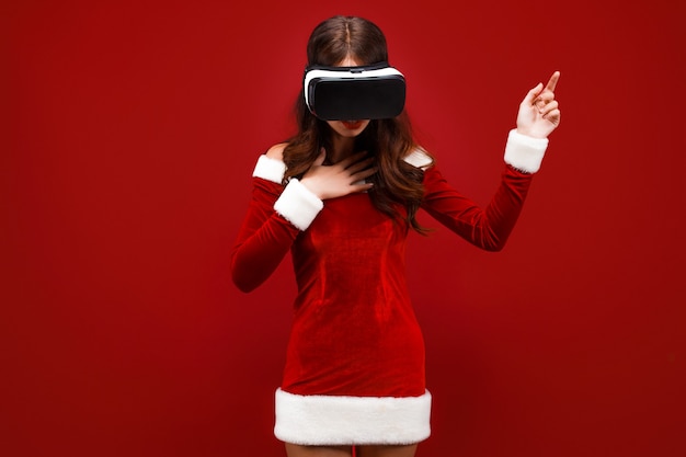 Verbaasd jong santameisje in kerstkleding die in hoofdtelefoon kijkt die handen opzij richt geïsoleerd op rode w...