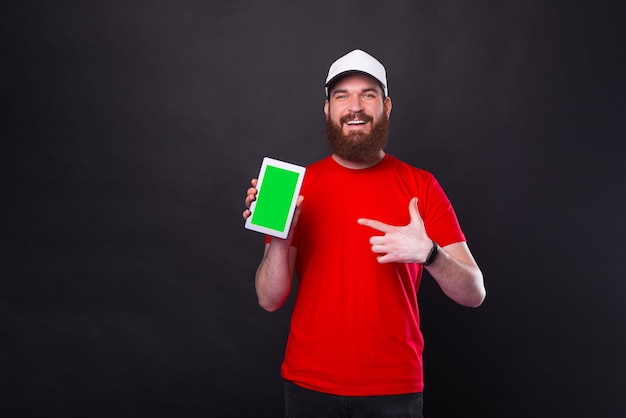 Verbaasd jonge bebaarde hipster man in rood t-shirt wijzend op groen scherm op tablet