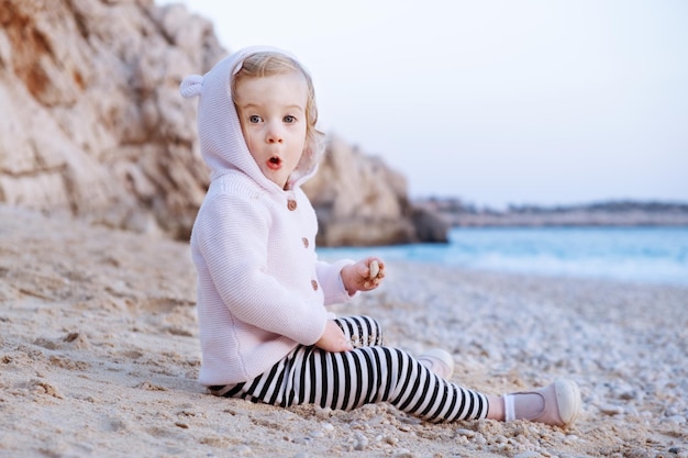 Verbaasd Kaukasisch babymeisje met open ogen zittend op het strand etend en spelend met zand aan de blauwe zee