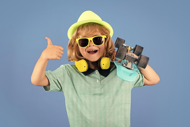 Verbaasd kind met duim omhoog Studio portret van stijlvol schattig jong kind jongen met skateboard geïsoleerd op blauwe achtergrond Modekind Stijlvol klein kind met een zomerkleding zonnebril en hoed